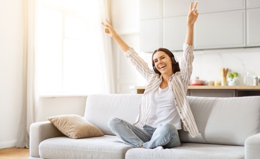 A cheerful woman wearing headphones sitting cross-legged on a couch with her arms raised in a peace sign, set in a bright living room.