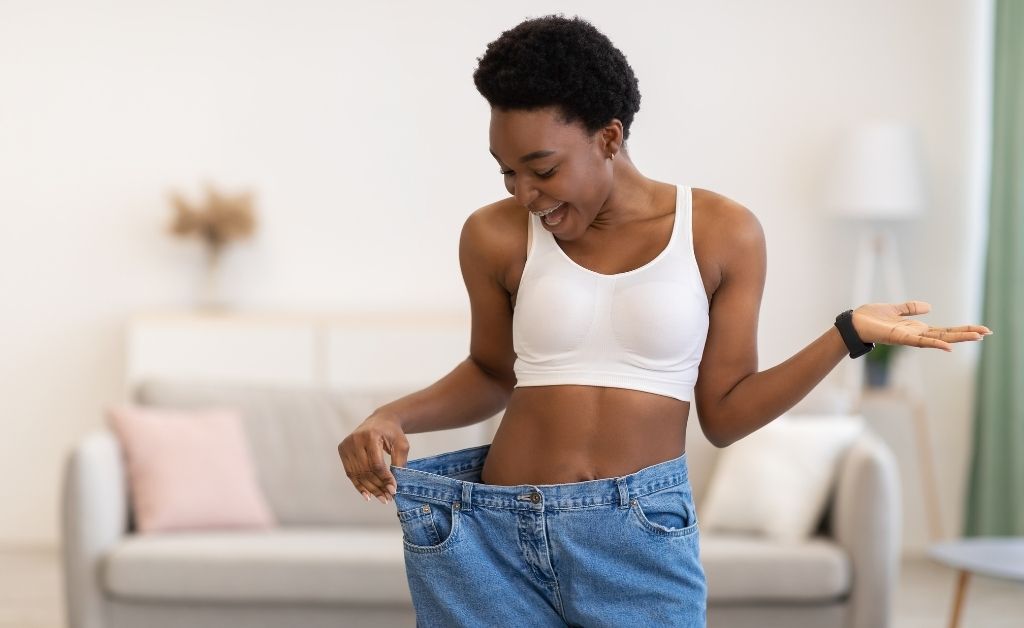 A joyful young woman with short, curly dark hair stands in a living room, smiling as she pulls out the waistband of a pair of oversized blue jeans to show her weight loss. She is wearing a white sports bra and a fitness tracker on her wrist. The background is a brightly lit, modern living room with a grey sofa and soft pink pillows.