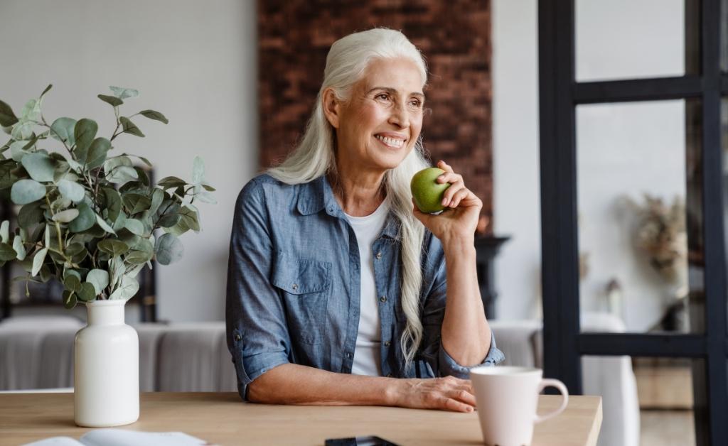 An older woman with long, white hair smiles warmly while sitting at a wooden table. She is wearing a blue button-down shirt and holds a green apple in her hand. On the table in front of her are a white mug and a potted eucalyptus plant. The background features a bright, modern interior with a brick accent wall and a black-framed window.