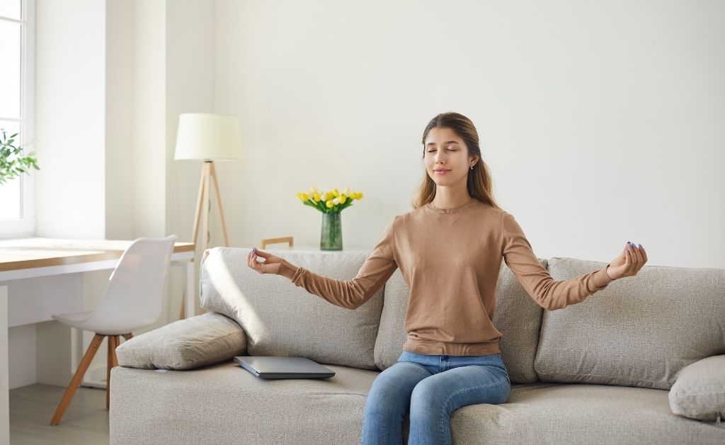 A young woman sits on a beige sofa in a bright, minimalist room, meditating with her eyes closed and hands in a mudra pose. A closed laptop sits beside her, suggesting a break from work.