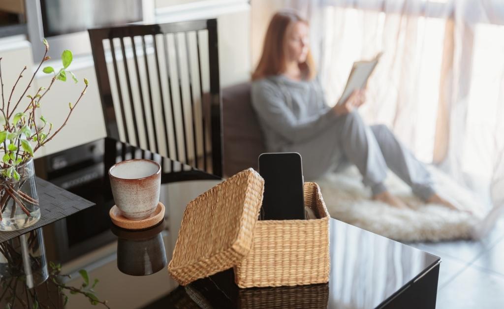A woman sits comfortably on a rug in the background, focused on reading a book, while her smartphone is tucked away in a small wicker basket on a table in the foreground. The image illustrates a successful effort to minimize digital distraction, with the phone physically separated from her personal space to allow for deep focus.